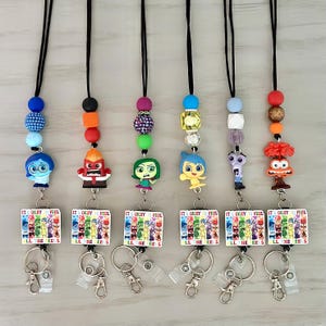 May include: A collection of lanyards featuring character charms and beaded accents. Each lanyard has a different character charm, including Anger, Sadness, Joy, and Fear. The lanyards also have a rectangular tag that reads "IT'S OKAY TO FEEL".