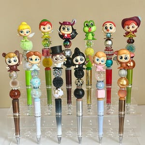 May include: A set of 14 colorful pens with various Disney character toppers. The pens are displayed on a clear acrylic stand. The toppers include characters like Peter Pan, Tinkerbell, Captain Hook, and more.