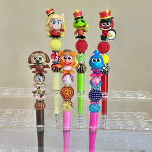 May include: A collection of colorful pens featuring various Muppet characters. Each pen has a unique design, with character figures atop beaded shafts in shades of pink, green, yellow, and red. The pens are displayed on a clear, tiered stand.
