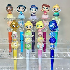 May include: A collection of decorative pens featuring fairy figurines. Each pen has a unique design with colorful accents, including orange, yellow, blue, and pink. The pens are displayed on a clear, tiered shelf.
