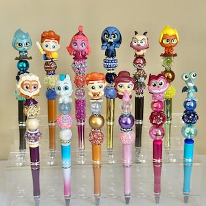 May include: A set of 12 colorful pens decorated with various cartoon characters. Each pen features a different character, including a blue dragon, a purple cat, a pink mermaid, and a yellow lion. The pens are all topped with a clear plastic bead and a small charm.