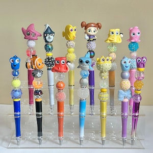 May include: A set of 14 colorful pens decorated with various characters from the Finding Nemo and Finding Dory movies. The pens are displayed on a clear acrylic stand.