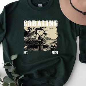 May include: Dark green sweatshirt with a graphic from the film "Coraline." The design features the word "CORALINE" above an illustration of the character and the text "EST. 2009."