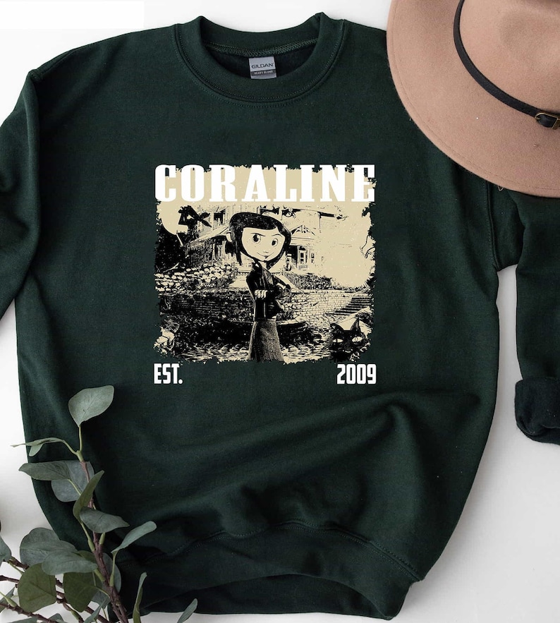 May include: Dark green sweatshirt with a graphic from the film "Coraline." The design features the word "CORALINE" above an illustration of the character and the text "EST. 2009."