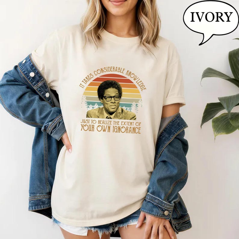 May include: Ivory-colored t-shirt featuring a retro design with a portrait of a man wearing glasses, set against a sunset backdrop. The text reads, "IT TAKES CONSIDERABLE KNOWLEDGE JUST TO REALIZE THE EXTENT OF YOUR OWN IGNORANCE."