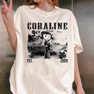May include: Cream-coloured t-shirt with a black and white graphic of a character from the film "Coraline" in front of a house. The word "CORALINE" is printed above the graphic, with "EST. 2009" below.