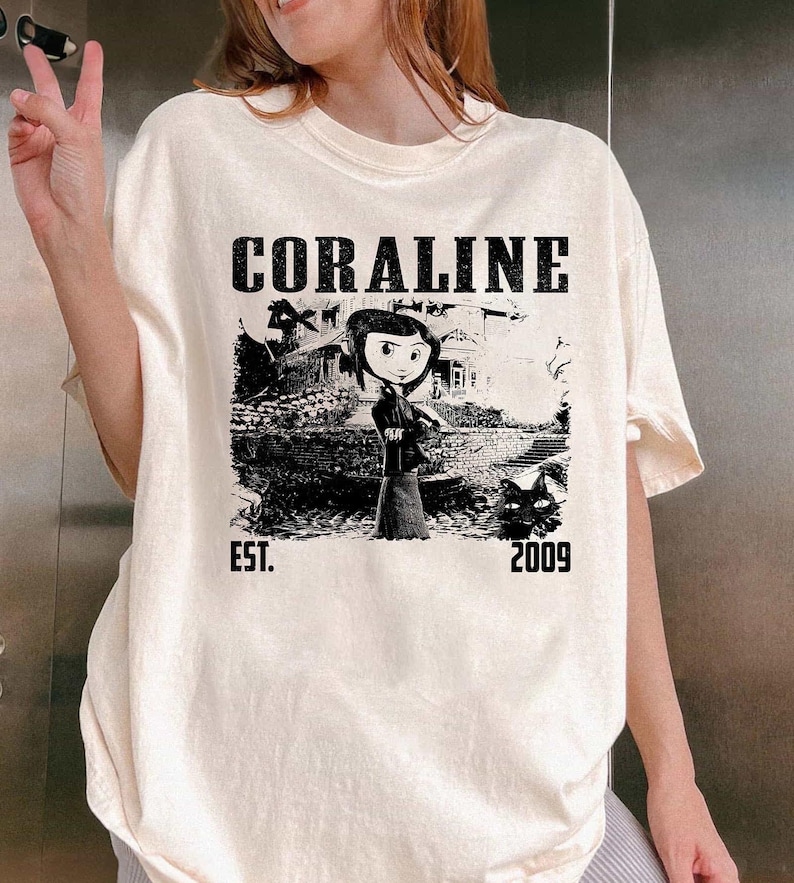 May include: Cream-coloured t-shirt with a black and white graphic of a character from the film "Coraline" in front of a house. The word "CORALINE" is printed above the graphic, with "EST. 2009" below.