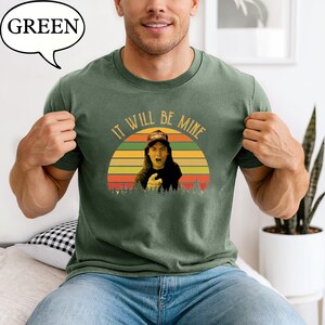 May include: Green t-shirt with a graphic of a person pointing, set against a sunset design. The text "IT WILL BE MINE" arches above the image. The shirt also has the word "GREEN" in a speech bubble.