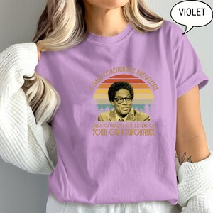 May include: Violet-colored t-shirt with a retro design featuring a portrait of a man wearing glasses, set against a sunset-colored background. The text reads, "It takes considerable knowledge just to realize the extent of your own ignorance."