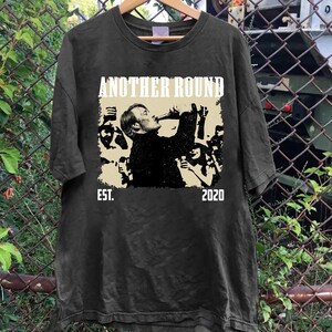 May include: Dark gray t-shirt with a graphic that reads "ANOTHER ROUND" above a black and white image of a person drinking. The text "EST. 2020" is below the image. The shirt is hanging on a wooden hanger.