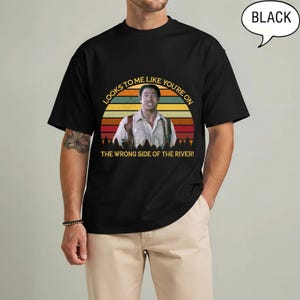 May include: Black t-shirt with a graphic featuring a man and the text "LOOKS TO ME LIKE YOU'RE ON THE WRONG SIDE OF THE RIVER!". The graphic is set against a sunset design. A speech bubble with the word "BLACK" is in the upper right corner.