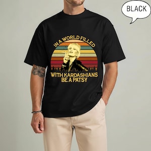 May include: Black t-shirt featuring a retro design with a woman holding a cigarette, set against a sunset backdrop. The text reads "IN A WORLD FILLED WITH KARDASHIANS BE A PATSY".