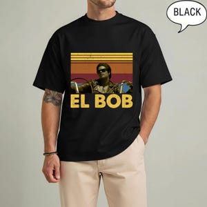 May include: Black t-shirt featuring a retro graphic of a person wearing sunglasses and a leather jacket, driving a vehicle. The text "EL BOB" is in yellow, with a sunset-style background. The word "BLACK" is in a speech bubble.