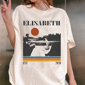 May include: Cream-colored t-shirt featuring the name "ELISABETH" in a bold, black font above a retro landscape design. The design includes a red sun, figures, and the text "EST. 2020" in a vintage style.