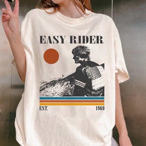 May include: Cream-colored t-shirt featuring the text "EASY RIDER" above a graphic of a person and a sunset. Below the graphic are the words "EST. 1969" and horizontal stripes in orange, blue, and brown.