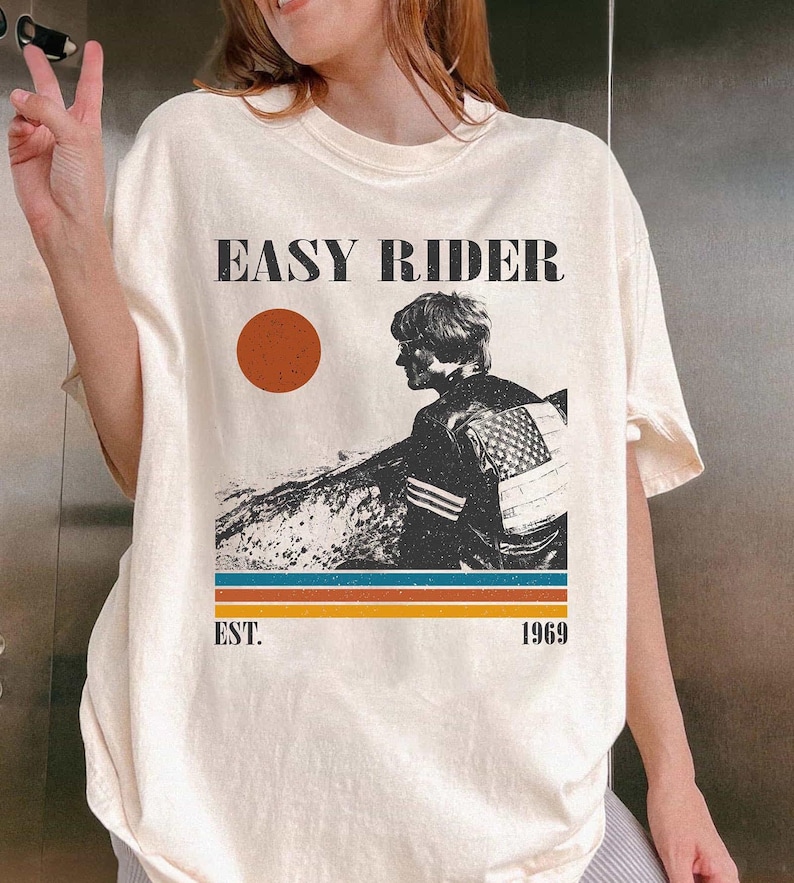 May include: Cream-colored t-shirt featuring the text "EASY RIDER" above a graphic of a person and a sunset. Below the graphic are the words "EST. 1969" and horizontal stripes in orange, blue, and brown.