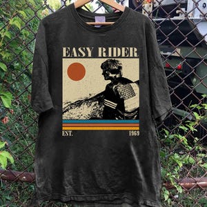 May include: Black t-shirt featuring a vintage graphic with the text "EASY RIDER" above an illustration of a person and a sun. Below the graphic are horizontal stripes in orange, blue, and brown, with "EST. 1969" below.