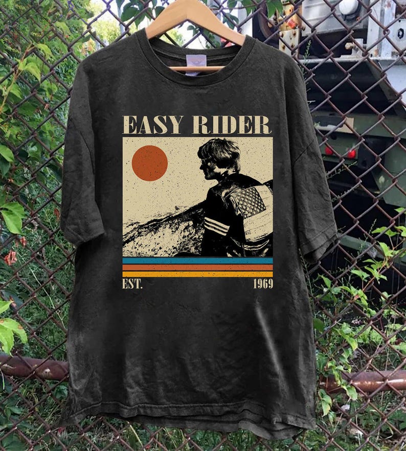 May include: Black t-shirt featuring a vintage graphic with the text "EASY RIDER" above an illustration of a person and a sun. Below the graphic are horizontal stripes in orange, blue, and brown, with "EST. 1969" below.