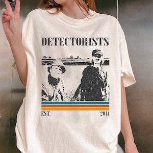 May include: Cream-colored t-shirt featuring a graphic of two people with the word "DETECTORISTS" above them. Below the image are the words "EST. 2014" in a vintage style. The shirt has a crew neck and short sleeves.