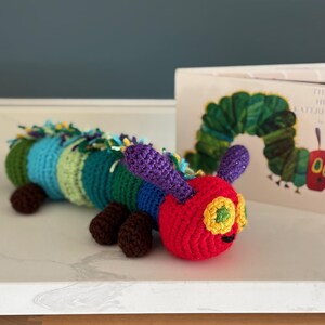 May include: A handmade crochet caterpillar toy in red, green, blue, and purple, with brown feet. The caterpillar is next to a copy of "The Very Hungry Caterpillar" book. The toy is approximately 25 cm long.
