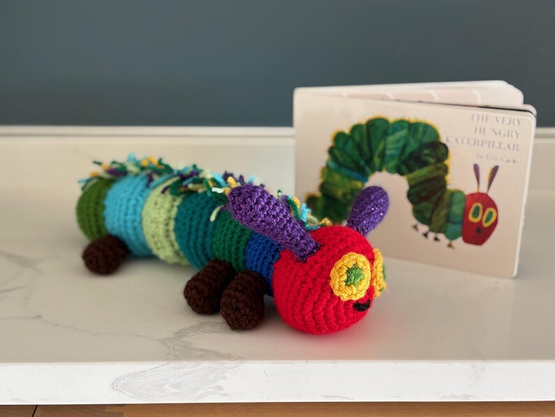 May include: A handmade crochet caterpillar toy in red, green, blue, and purple, with brown feet. The caterpillar is next to a copy of "The Very Hungry Caterpillar" book. The toy is approximately 25 cm long.