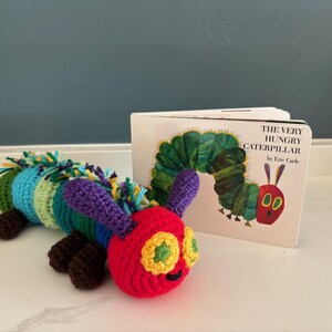 May include: A colourful crocheted caterpillar toy, with segments of red, green, blue, and purple, beside a copy of "The Very Hungry Caterpillar" book. The caterpillar has yellow eyes and brown feet. The book cover shows a green caterpillar.