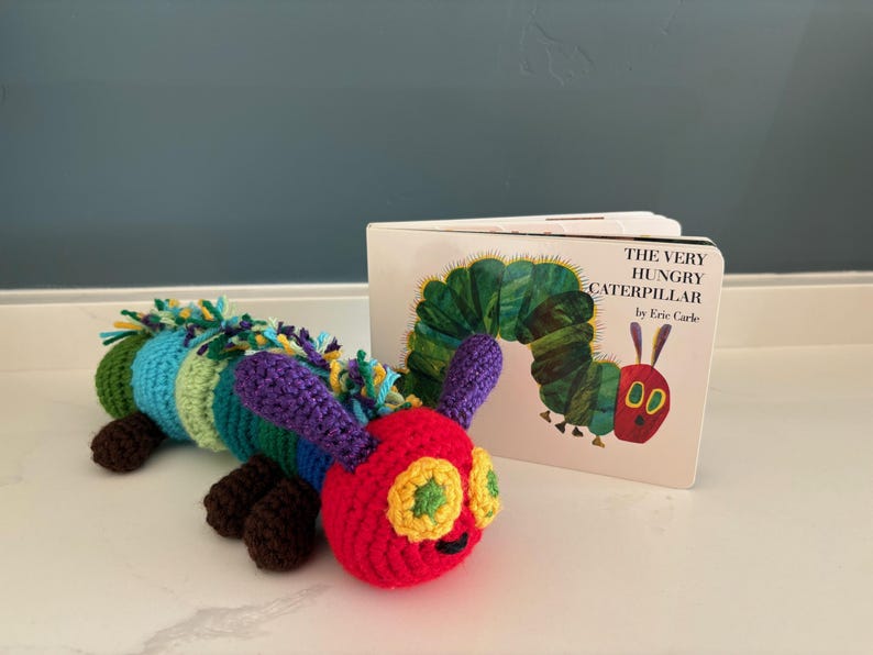 May include: A colourful crocheted caterpillar toy, with segments of red, green, blue, and purple, beside a copy of "The Very Hungry Caterpillar" book. The caterpillar has yellow eyes and brown feet. The book cover shows a green caterpillar.