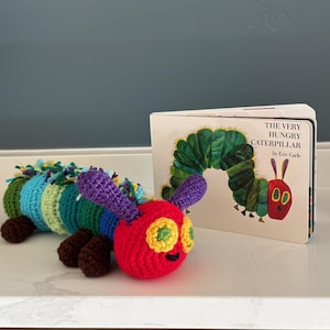May include: A vibrant, crocheted caterpillar toy with a red head, green and yellow eyes, and a multi-coloured body. Next to it is a copy of "The Very Hungry Caterpillar" book by Eric Carle.