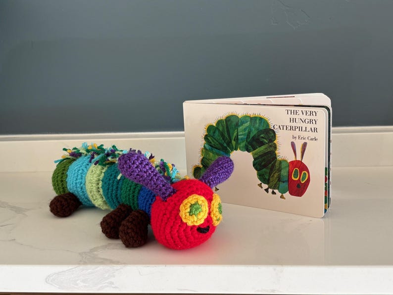 May include: A vibrant, crocheted caterpillar toy with a red head, green and yellow eyes, and a multi-coloured body. Next to it is a copy of "The Very Hungry Caterpillar" book by Eric Carle.