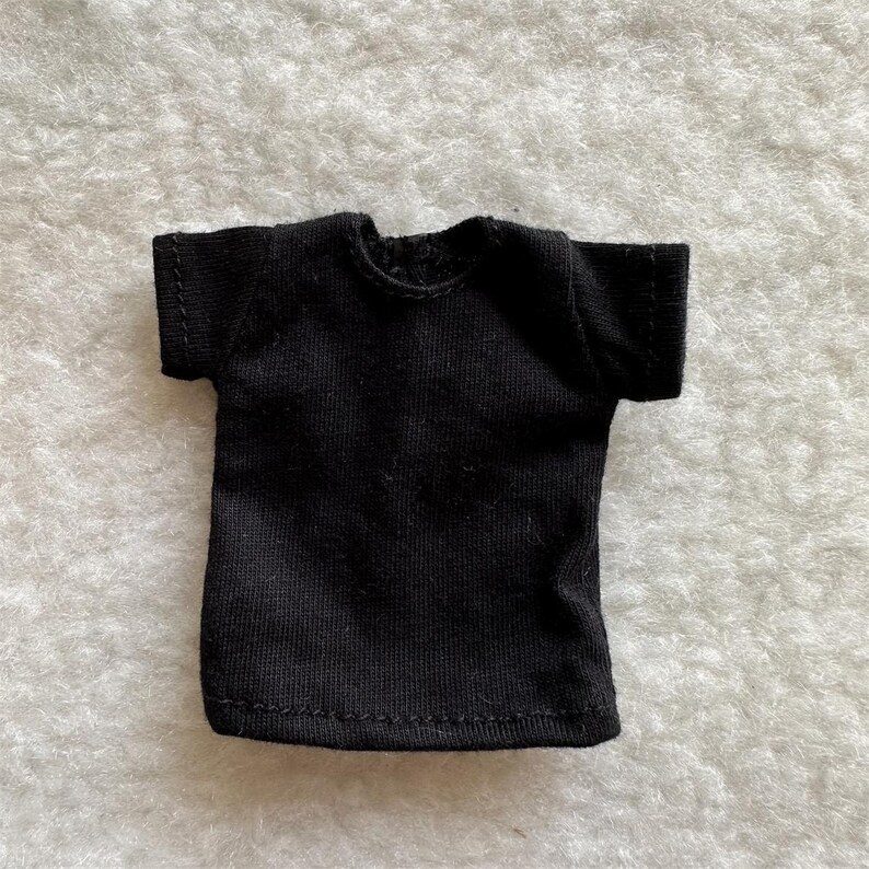 Puede incluir: Una peque&ntilde;a camiseta negra lisa con mangas cortas. Tiene cuello redondo. La tela parece ser un material suave y texturizado. Esta prenda es probablemente para una mu&ntilde;eca u otra figura peque&ntilde;a.