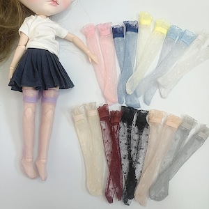 May include: A doll in a white t-shirt and navy skirt, with an assortment of lace socks. The socks are in shades of pink, blue, yellow, and white, and feature a polka dot pattern. The socks are designed for dolls.