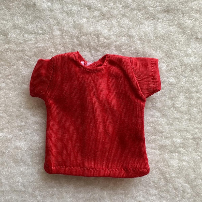 Puede incluir: Una peque&ntilde;a camiseta roja lisa con mangas cortas. La camiseta est&aacute; hecha de tejido de punto y tiene un cuello redondo simple. La prenda es probablemente para una mu&ntilde;eca o un juguete peque&ntilde;o.