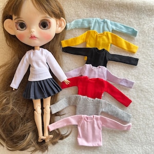 Puede incluir: Una mu&ntilde;eca con una camisa blanca de manga larga y una falda plisada gris. Se muestran varias camisetas de manga larga en miniatura en varios colores: azul claro, amarillo, negro, lavanda, rojo, gris y rosa. Las camisetas son para mu&ntilde;ecas.