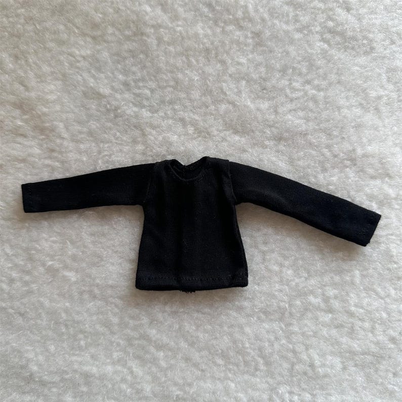 Puede incluir: Una peque&ntilde;a camisa negra de manga larga. La camisa est&aacute; hecha de una tela suave y texturizada y tiene cuello redondo. Las mangas son largas y se extienden a los lados. La camisa est&aacute; dise&ntilde;ada para una mu&ntilde;eca.