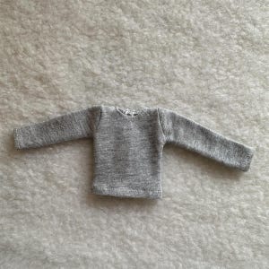 Puede incluir: Una peque&ntilde;a camiseta de manga larga de color gris claro. La prenda tiene cuello redondo y est&aacute; hecha de una tela suave y texturizada. Las mangas son largas y llegan hasta las mu&ntilde;ecas. La camiseta es probablemente para una mu&ntilde;eca o un ni&ntilde;o peque&ntilde;o.