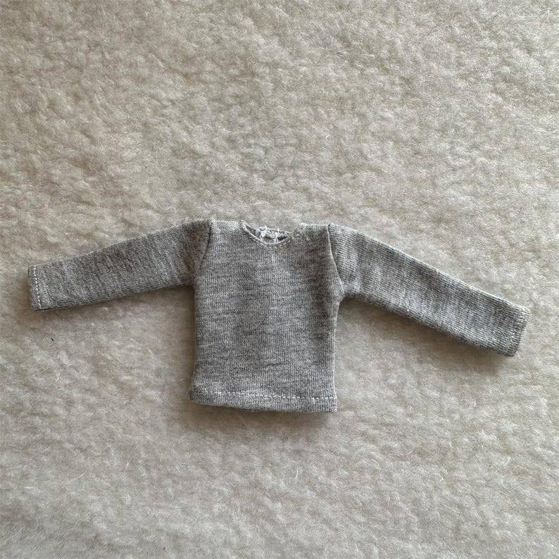Puede incluir: Una peque&ntilde;a camiseta de manga larga de color gris claro. La prenda tiene cuello redondo y est&aacute; hecha de una tela suave y texturizada. Las mangas son largas y llegan hasta las mu&ntilde;ecas. La camiseta es probablemente para una mu&ntilde;eca o un ni&ntilde;o peque&ntilde;o.