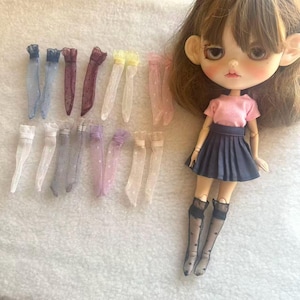 Pode incluir: Uma boneca vestindo uma camiseta rosa e uma saia plissada azul marinho, com uma coleção de meias transparentes para bonecas em várias cores. As meias são em tons de azul, roxo, amarelo e rosa, com acabamentos em renda.