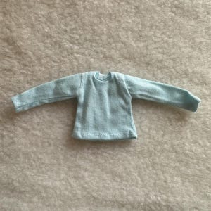 Puede incluir: Una camiseta de manga larga azul claro. La camiseta est&aacute; hecha de un material suave y tejido. Las mangas son largas y se extienden hacia los lados. El cuello es redondo simple. La camiseta est&aacute; dise&ntilde;ada para una mu&ntilde;eca.