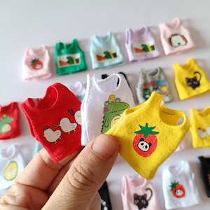 May include: A selection of small doll tank tops in diverse colours, including red, white, yellow, green, and pink. Each top has a unique graphic, like a strawberry, cat, or cartoon animal. The tops are held by a hand.