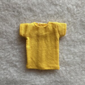 Puede incluir: Una peque&ntilde;a camiseta amarilla brillante con mangas cortas. La camiseta est&aacute; hecha de una tela suave y tiene un dise&ntilde;o simple y cl&aacute;sico. Las costuras son visibles y la camiseta parece hecha a mano. Esta prenda en miniatura es probablemente para una mu&ntilde;eca u otra figura peque&ntilde;a.