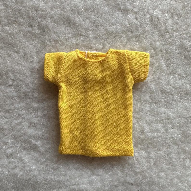 Puede incluir: Una peque&ntilde;a camiseta amarilla brillante con mangas cortas. La camiseta est&aacute; hecha de una tela suave y tiene un dise&ntilde;o simple y cl&aacute;sico. Las costuras son visibles y la camiseta parece hecha a mano. Esta prenda en miniatura es probablemente para una mu&ntilde;eca u otra figura peque&ntilde;a.