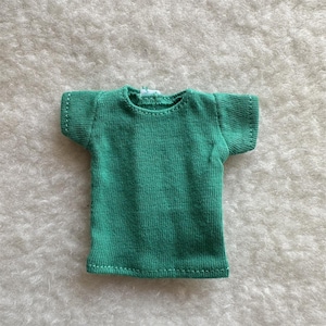 Puede incluir: Una peque&ntilde;a camiseta verde esmeralda con mangas cortas. La camiseta est&aacute; hecha de una tela de punto y tiene costuras blancas a lo largo de las costuras. La camiseta se muestra sobre una superficie blanca texturizada.