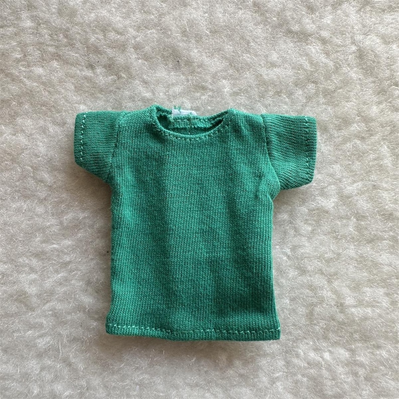 Puede incluir: Una peque&ntilde;a camiseta verde esmeralda con mangas cortas. La camiseta est&aacute; hecha de una tela de punto y tiene costuras blancas a lo largo de las costuras. La camiseta se muestra sobre una superficie blanca texturizada.