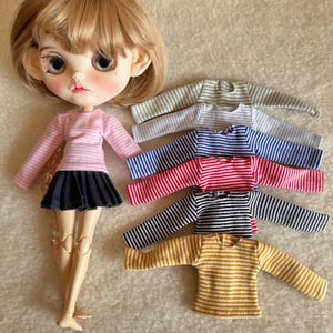 May include: A doll wearing a pink and white striped long-sleeve shirt and a black skirt. Several other striped long-sleeve shirts in various colors, including green, blue, red, black, and yellow, are displayed nearby. The shirts are doll-sized.