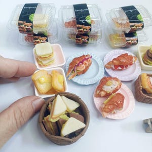 May include: A collection of miniature food items, including sandwiches, croissants, and toast. Some are in clear containers labelled "Sandwich". Others are on small plates and in a woven basket. The food items are arranged on a white surface.