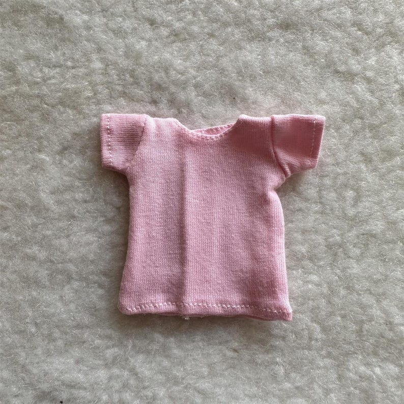 Puede incluir: Una peque&ntilde;a camiseta rosa claro con mangas cortas. La camiseta est&aacute; hecha de una tela suave y tiene un dise&ntilde;o simple y cl&aacute;sico. Las costuras son visibles, agregando un toque artesanal.