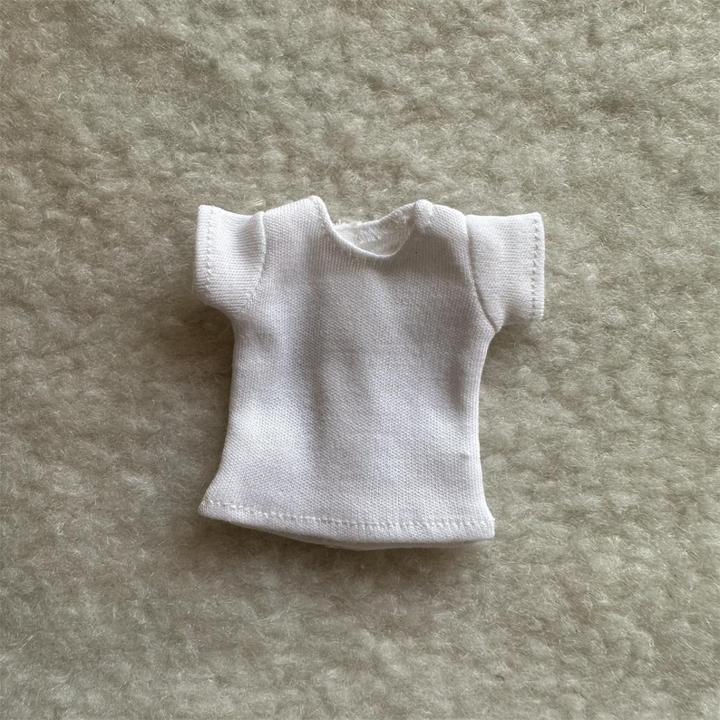 Puede incluir: Una peque&ntilde;a camiseta blanca con mangas cortas. La camiseta est&aacute; hecha de una tela suave y texturizada y tiene un cuello redondo. Las costuras son visibles y la camiseta parece nueva.