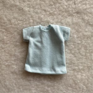 Puede incluir: Una camiseta azul claro con mangas cortas. La camiseta est&aacute; hecha de una tela suave y texturizada y tiene un dise&ntilde;o simple y cl&aacute;sico. La camiseta es probablemente para una mu&ntilde;eca o un juguete peque&ntilde;o.