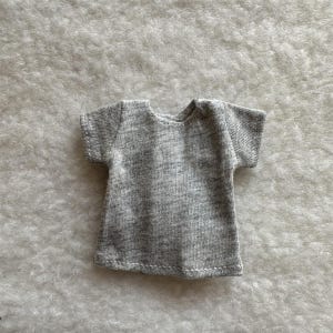 Puede incluir: Una peque&ntilde;a camiseta gris con mangas cortas. La camiseta est&aacute; hecha de una tela suave y jaspeada y tiene un dise&ntilde;o simple y cl&aacute;sico. El cuello es redondo y las mangas son cortas y est&aacute;n terminadas con un dobladillo simple.