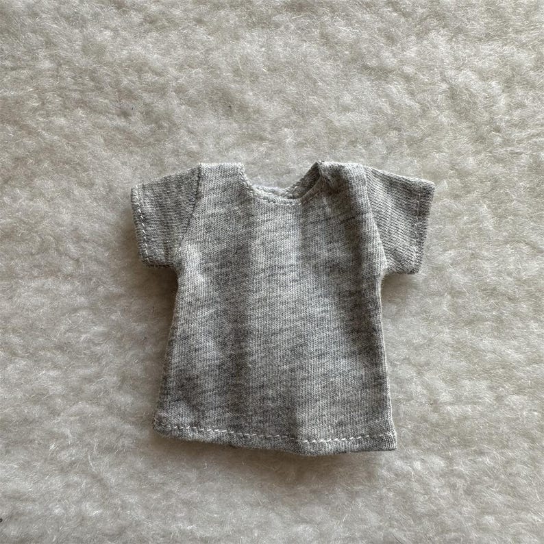 Puede incluir: Una peque&ntilde;a camiseta gris con mangas cortas. La camiseta est&aacute; hecha de una tela suave y jaspeada y tiene un dise&ntilde;o simple y cl&aacute;sico. El cuello es redondo y las mangas son cortas y est&aacute;n terminadas con un dobladillo simple.